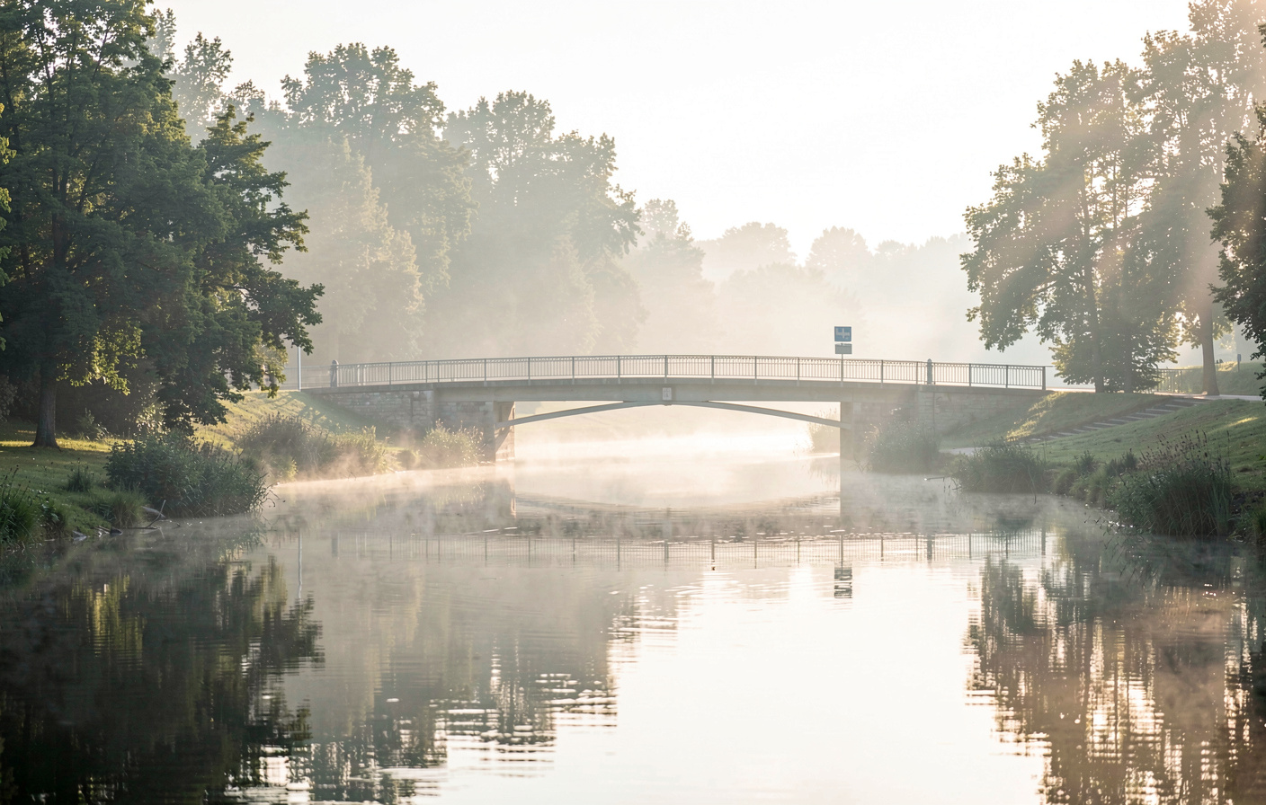 En bro over Fuldafloden nær Eichenzell tidligt om morgenen, blød sol gennem let tåge, vandet spejler træerne, bredt landskabsbillede.