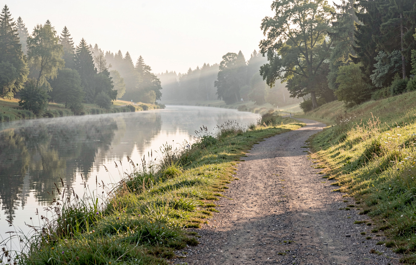 En sti langs floden Fulda ved Eichenzell tidligt om morgenen, vandet spejler træerne, blød tåge over engene, vidvinkel fra lav position med stien som ledende linje ind mod skovbrynet.